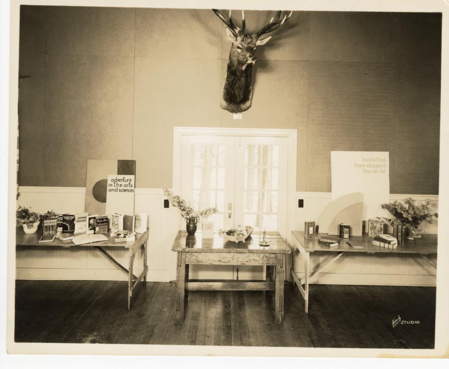 Black-and-white photo of Stevenson Community Library interior, with a deer head mounted on the wall, various items displayed on tables, and a wood floor 